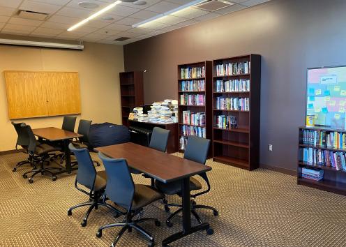 two brown tables with four office chairs at each occupy the foreground of the image with a few bookshelves in the background