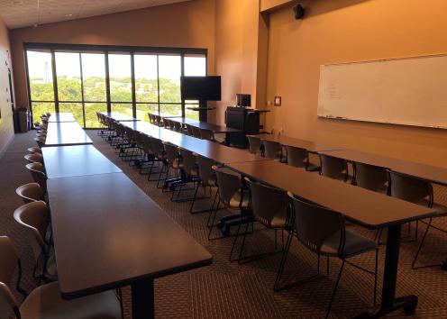 desks with chairs face a whiteboard, podium, and TV to the right of the room with bright light coming in from the windows in the background