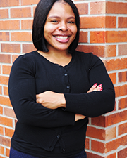 Woman with short, black hair wearing black button-up sweater leans against brick wall smiling at the camera.